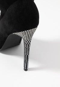 Musta nubukista valmistettu korkeakorkoinen kenkä, jossa on stiletto-korko, joka on koristeltu valkoisilla strasseilla, korostaen kimaltelevaa pintarakennetta jaElegantti muotoilu.