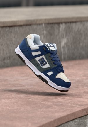 Sinine ja valge DC rulluiskade king tumesiniste paelte, valge tallaga ja logoga, hõljumas roosa betoonserva kohal.