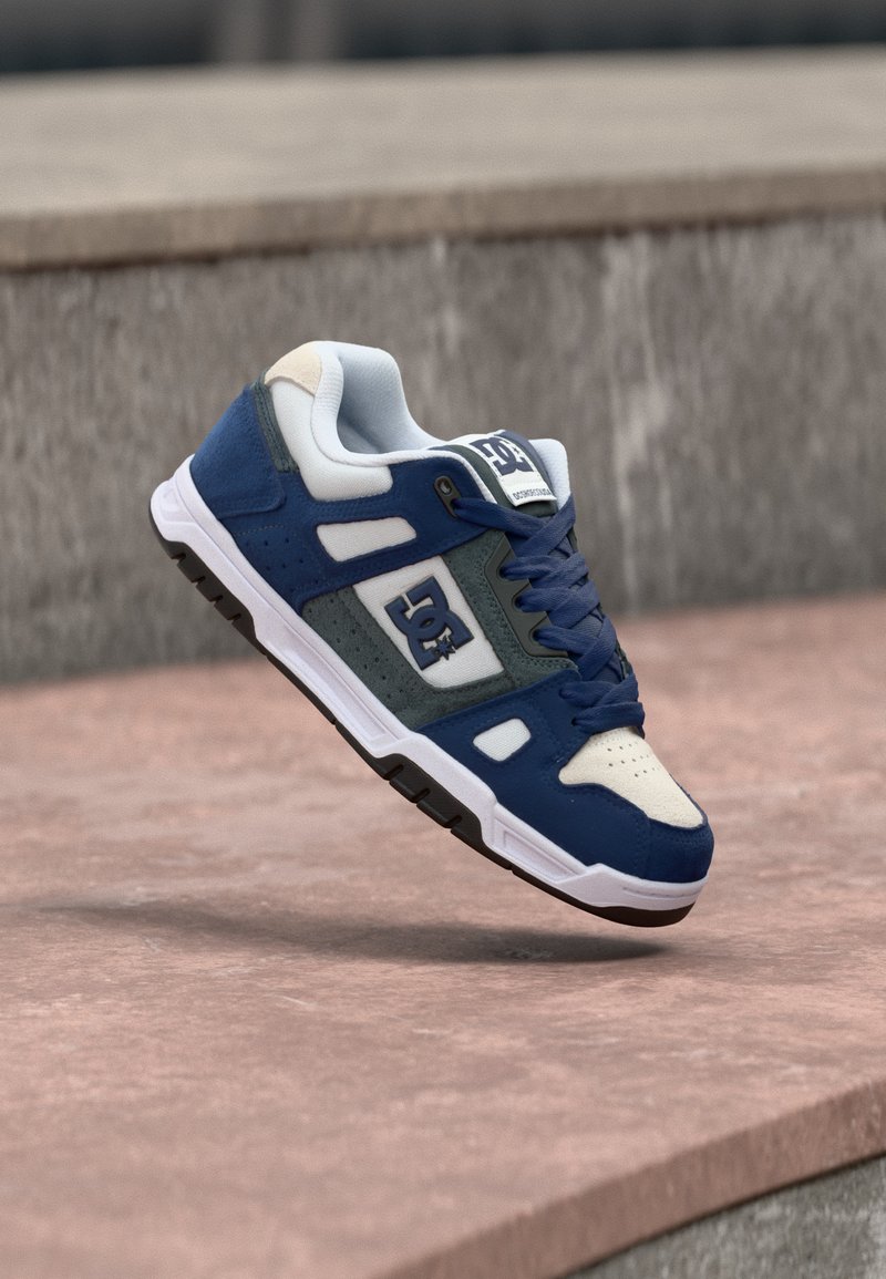 Μπλε και λευκό παπούτσι DC για σκέιτ με σκουρομπλε κορδόνια, λευκή σόλα και λογότυπο, να αιωρείται πάνω από μια ροζ σκυρόδετη άκρη.