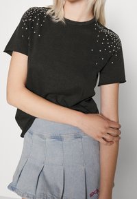 Schwarzes T-Shirt mit kurzen Ärmeln und verstreuten Strasssteinverzierungen auf den Schultern. Kombiniert mit einem hellen, plissierten Jeansrock.