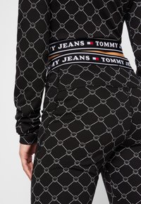 Tessuto nero con motivi a faccina sorridente bianca disposti a griglia. Fascia in vita marchiata Tommy Jeans con colori a contrasto e dettagli del logo.