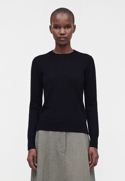 Femme aux cheveux courts portant un haut noir à manches longues et une jupe grise, debout devant un fond blanc uni.