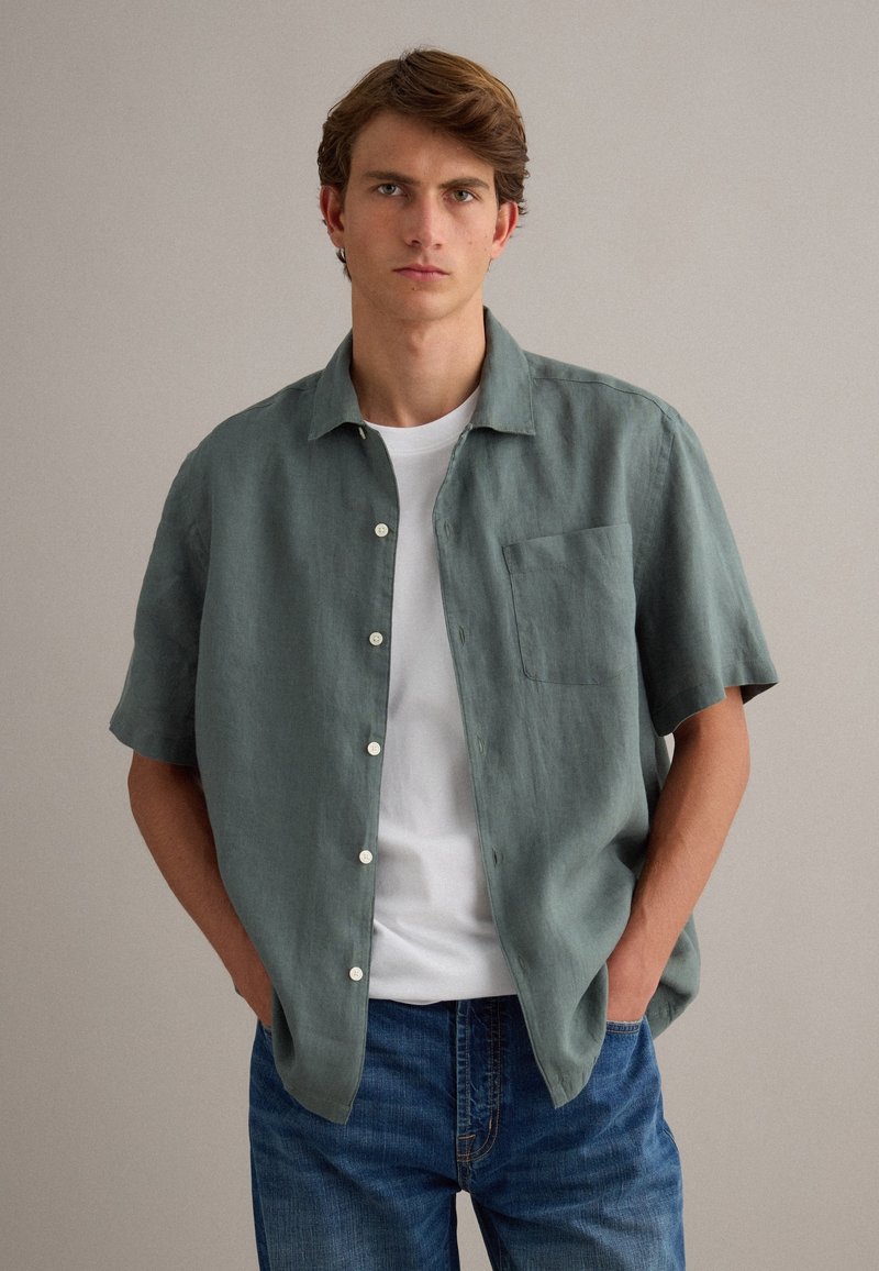 Joven con cabello castaño que lleva una camisa de manga corta verde abotonada sobre una camiseta blanca y jeans azules, con un fondo liso.