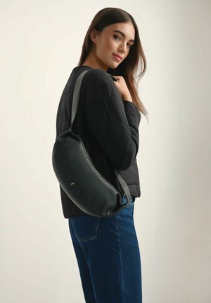 Młoda kobieta ubrana w czarną kurtkę i niebieskie dżinsy, nosząca czarną skórzaną torbę na ramię, patrząca przez ramię za siebie.