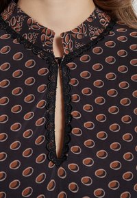 Svart tygblus med brunt cirkulärt mönster, har en scallopad svart spetskrage och en nyckelhålsöppning vid kragen.