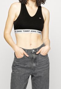 Bralette nera in cotone con scollatura a V profonda, caratterizzata da una fascia bianca con la scritta "TOMMY JEANS". Abbinato a jeans in denim grigio a vita alta.
