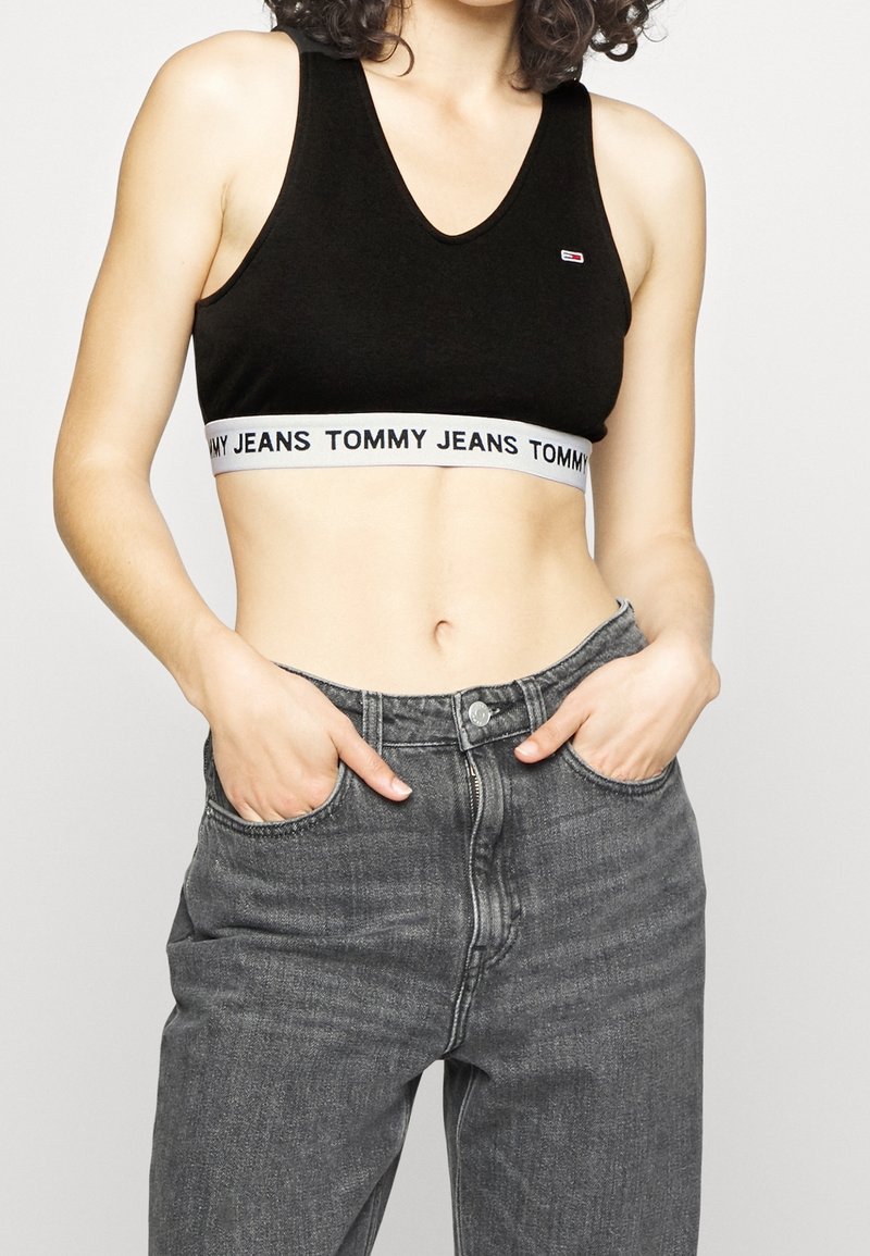 Bralette nera in cotone con scollatura a V profonda, caratterizzata da una fascia bianca con la scritta "TOMMY JEANS". Abbinato a jeans in denim grigio a vita alta.