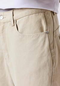 Détail de la poche d'un pantalon beige texturé avec bouton et passants pour ceinture, porté avec un haut gris clair rentré à l'intérieur.