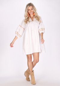 Rochie albă cu mâneci bufante și broderie florală pe manșete. Prezintă un șnur cu franjuri în față și o croială lejeră. Este purtată cu cizme cu toc de culoare bej.