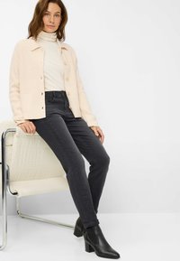 Cardigan lavorato a maglia di un leggero beige con bottoni, abbinato a jeans neri a vita alta e stivaletti neri con tacco, seduto su una sedia bianca.
