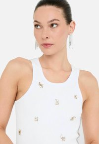 Een jonge vrouw met een strakke bun draagt een mouwloos wit topje versierd met decoratieve metalen elementen, wat een stijlvolle, moderne uitstraling biedt.
