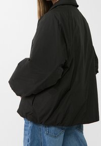 Giacca nera oversized realizzata in materiale impermeabile, dotata di colletto e orlo elastico, mostrata di lato con jeans in denim blu.