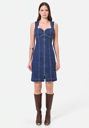 Femme portant une robe en denim bleu sans manches avec une fermeture éclair devant et des bottes marron jusqu'aux genoux, debout devant un fond clair uni.