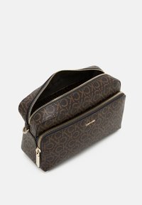 Brązowa saszetka Calvin Klein z powtarzającym się wzorem logo CK, dwiema zamykanymi na zamek kieszeniami oraz złotym logo Calvin Klein na przedniej kieszeni.