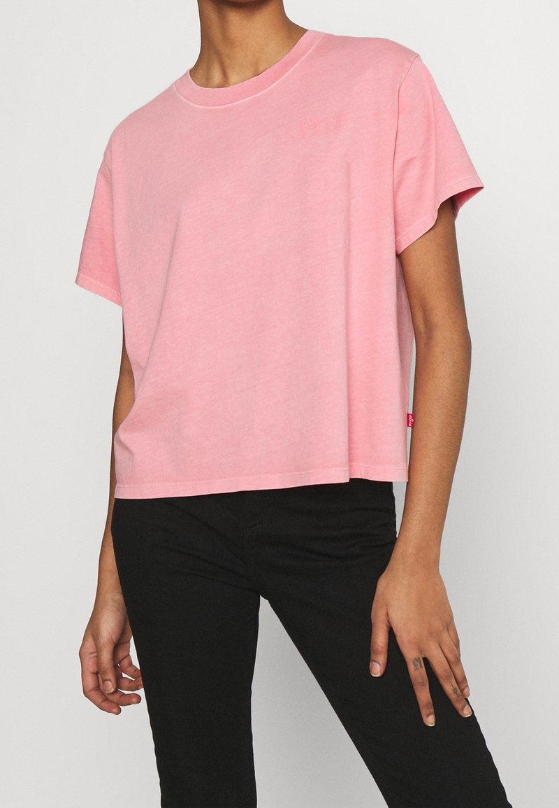 Personne portant un t-shirt rose ample à manches courtes et un pantalon noir, debout avec une main détendue le long du corps.