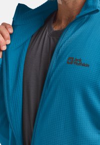 Veste légère teal avec une texture matelassée, fermeture éclair et un logo noir ; portée par-dessus une chemise gris foncé, mettant en valeur un design en couches.