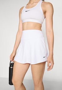 Conjunto de sujetador deportivo y falda en blanco, con un logotipo negro de Nike, textura de tela suave y una cinturilla ajustada. Botella de agua en la mano.
