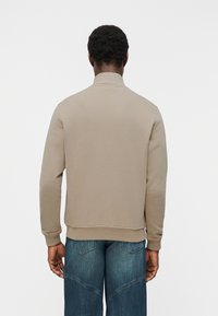 Άνδρας που φοράει μπεζ μπλούζα με μακριά μανίκια και μπλε τζιν, με την πλάτη του προς την κάμερα, πάνω σε απλό λευκό φόντο.