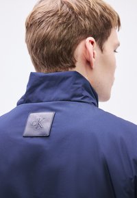 Ναυτικό μπουφάν με ψηλό γιακά και υφή υφάσματος. Διακριτικό λογότυπο cK σε τετράγωνο έμβλημα στην πλάτη. Λεπτό φινίρισμα.