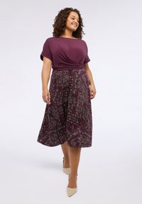 Femeie mergând cu încredere, îmbrăcată cu un top mov cu față răsucită și o fustă midi cu un model, purtând pantofi cu vârf ascuțit bej.