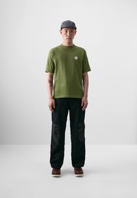 Grøn kortærmet t-shirt med et hvidt stjernesymbol, parret med sorte bukser med buede lapper og brune sko. Enkel, moderne design.