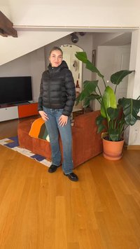 Žena v černé péřové bundě, džínách a černých botách stojí v obývacím pokoji s oranžovou pohovkou, velkou zelenou rostlinou a televizí na skříňce.