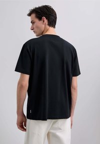 Tricou negru din bumbac, cu mâneci scurte, croială lejeră și decolteu rotund. Viziunea din spate arată o textură netedă și un detaliu subtil cu slit lateral.