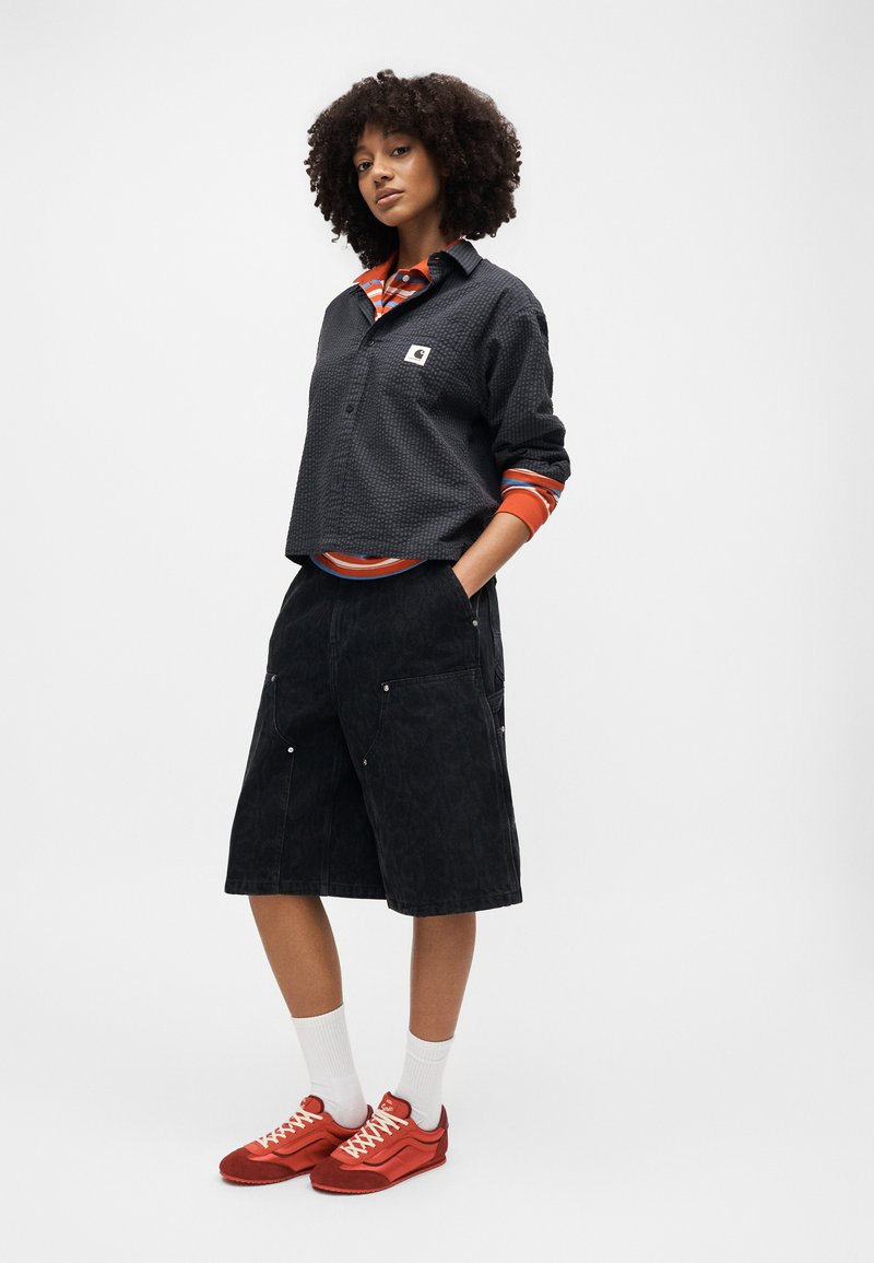 Frau mit lockigem Haar trägt ein schwarzes strukturiertes Hemd über einem orange gestreiften Top, schwarze knielange Shorts, weiße Socken und rote Turnschuhe.