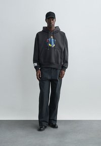 Graues, übergroßes Hoodie mit grafischem Katzendesign auf der Vorderseite, kombiniert mit weiten schwarzen Hosen. Schwarze Mütze mit Textelement.