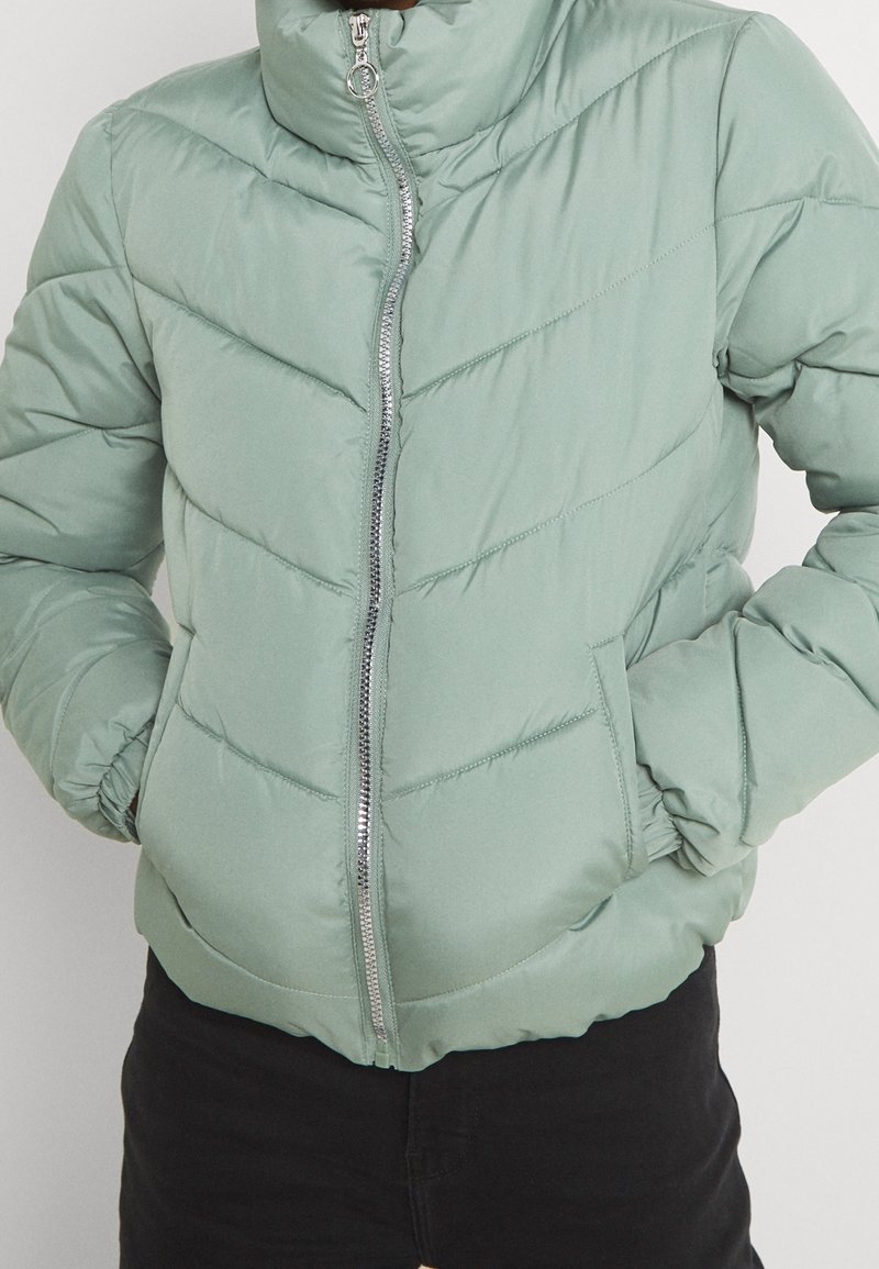 Veste matelassée vert clair avec un col montant, des coutures en chevron, une fermeture éclair argentée sur le devant et des poignets élastiques. Dispose de poches latérales et d'une texture lisse.