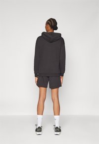Zwarte katoenen hoodie met een capuchon, gecombineerd met bijpassende donkere shorts. Model toont een ontspannen pasvorm, ribbels aan de manchetten en sportieve sneakers.