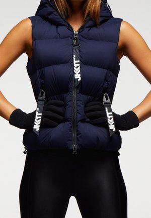 Kobieta w bezrękawniku pikowanym w kolorze granatowym z dużymi suwakami z logo i czarnych rękawiczkach, pozująca z rękami na biodrach na jednolitym tle.