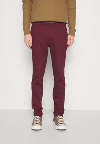 Chinos burdeos de corte slim con cierre de botón, que cuentan con bolsillos laterales y una suave tela de algodón. Combinados con zapatillas marrones.