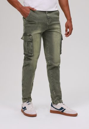 Pantalons cargo verts à coupe slim, avec plusieurs poches latérales, une texture délavée et un éclat subtil. Associés à des baskets blanches.