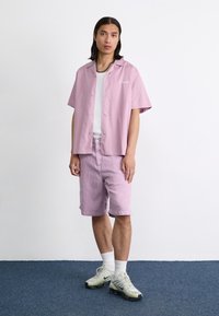 Korte mouwen, lichtroze button-up shirt, grijze tanktop eronder, bijpassende roze shorts en grijze sneakers met groene accenten. Eenvoudig ontwerp.