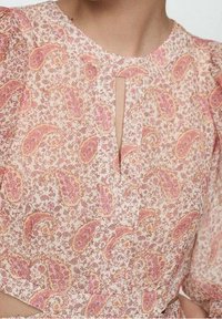 Detailní záběr na blůzu s paisley vzorem v růžové a béžové barvě, s výstřihem ve tvaru klíče a nařasenými rukávy, kterou má na sobě osoba.