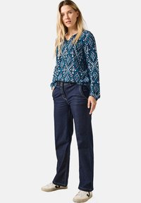 Blaue, gemusterte Bluse mit langen Ärmeln und einem Diamantdesign, kombiniert mit dunklen Jeans und weißen Sneakers. Lange, gerade geschnittene Hosen im Casual-Stil.