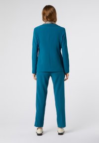 Costume bleu sarcelle avec un blazer ajusté et un pantalon droit. Présente un motif décoratif sur le col. Le tissu semble lisse avec un design structuré.