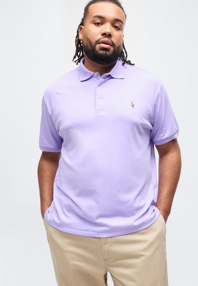 Άνδρας με ντραντλοκς που φοράει ανοιχτό μωβ πόλο μπλουζάκι και μπεζ παντελόνι, στέκεται με τα χέρια στις τσέπες μπροστά σε απλή φόντο.