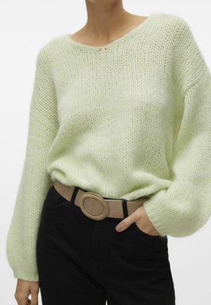 Personne portant un pull tricoté vert clair avec des manches ballon, un pantalon noir et une ceinture beige avec une boucle ovale, main dans la poche.