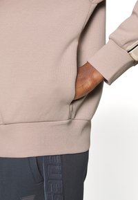 Sweatshirt beige avec des poignets et un ourlet côtelés, comportant une poche latérale. Assorti à un pantalon de survêtement bleu foncé, affichant une marque texturée.