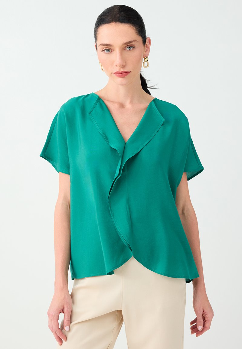 Femme aux cheveux foncés en queue de cheval porte un chemisier vert à manches courtes avec détail volanté et un pantalon écru sur un fond clair uni.