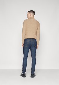 Bežinė glotniai megzta turtlenkio megztinis, dėvimas su tamsiai mėlynomis tiesiomis kelnėmis ir juodais batais, matomas iš nugaros, prieš baltą fono.