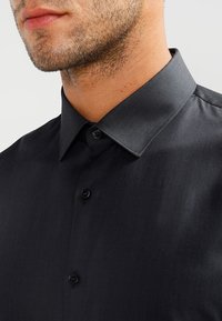 Black button-up shirt featuring a stiff collar, smooth texture, and subtle sheen. The shirt has a slim fit with black buttons.