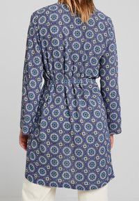 Långärmad, bälteklädd kappa i blått och vitt mönstrat tyg, med cirkulära motiv. Materialet verkar mjukt med en avslappnad passform.