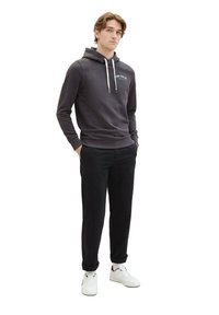 Felpa grigia scura con tasca a canguro, cappuccio con cordino e logo sul petto, abbinata a pantaloni neri e sneakers bianche.