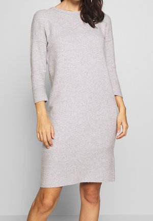 Frau trägt ein hellgraues, knielanges, langärmliges Strickkleid vor schlichter Hintergrund.