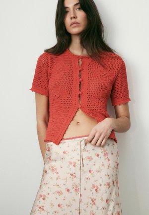 Junge Frau trägt ein rotes, gehäkeltes Kurzarmshirt mit Knöpfen und einen weißen Blumenrock, steht vor einem einfarbigen Hintergrund.