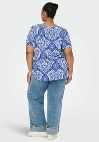 Kvinna med uppsatt hår, iklädd en blå och vit mönstrad kortärmad topp, lösa blå jeans med uppvikta ärmar och vita sneakers, som står mot en enfärgad bakgrund.