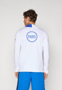 Long-sleeve white shirt with blue collar; round blue logo reading "PARIS" on back. Worn with blue shorts. Smooth, lightweight fabric.